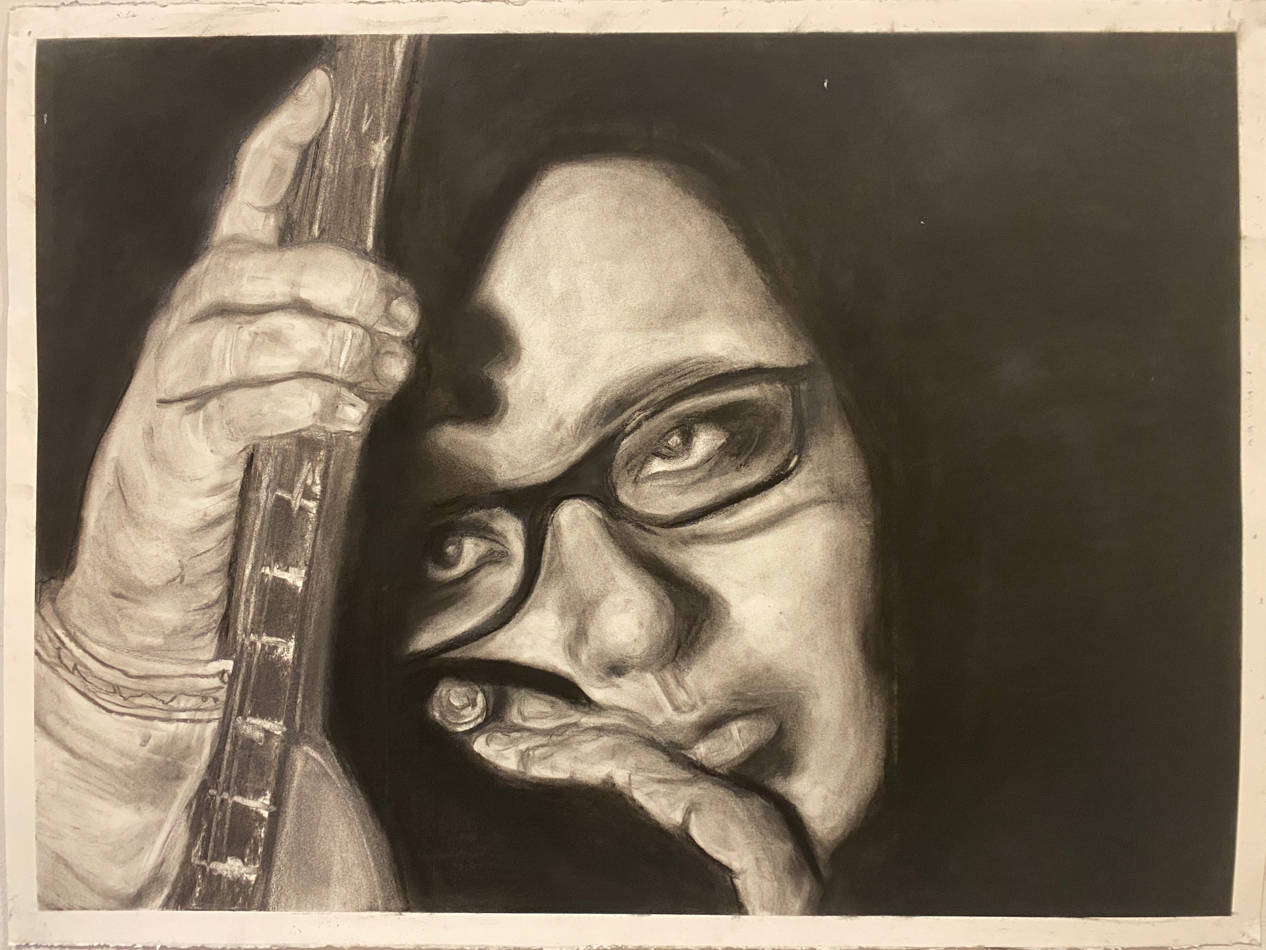 Image ID: A dark charcoal drawing focused on a pale person's face wearing glasses and holding a guitar with a forlorn expression.