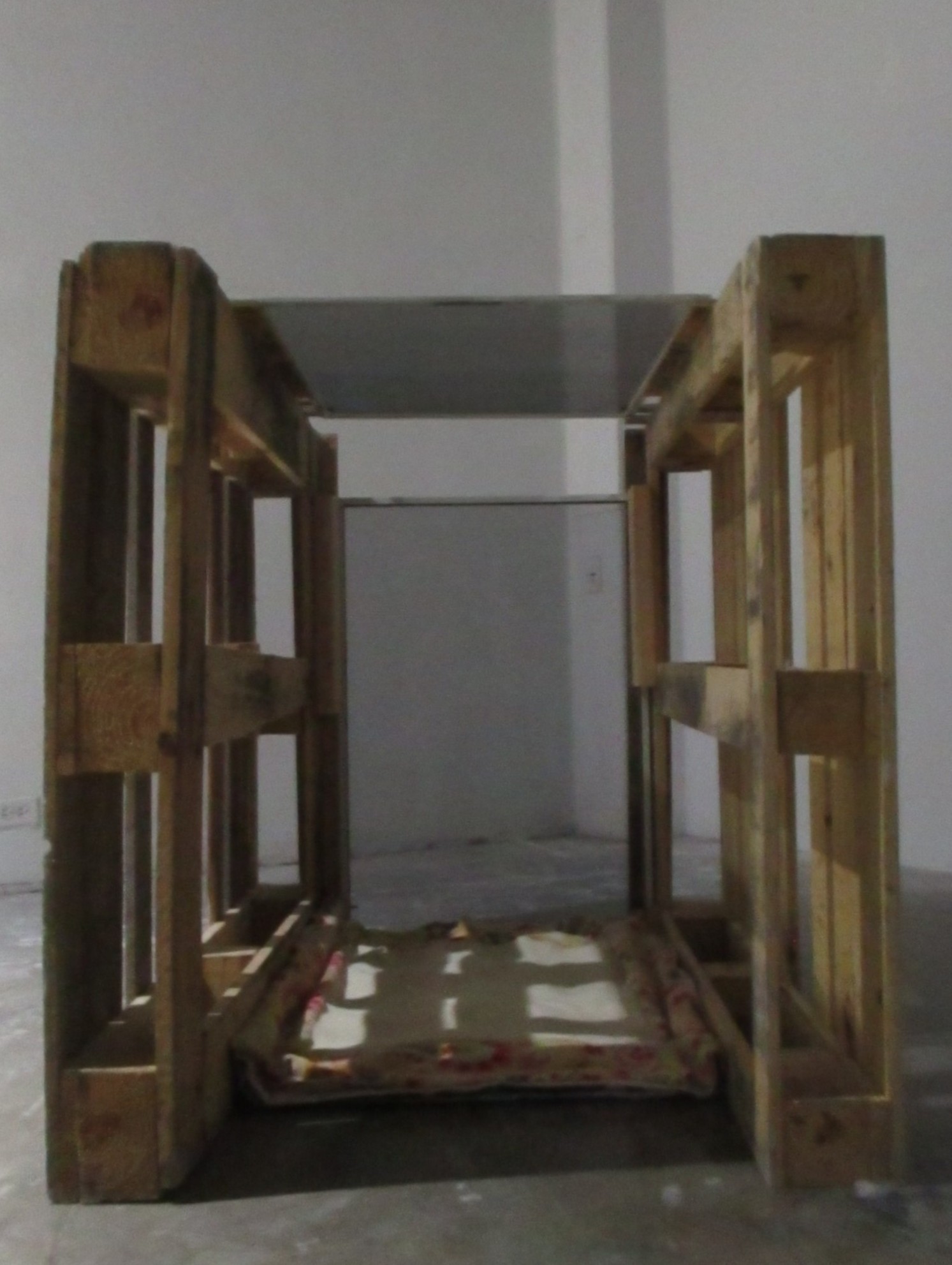 Image ID: A close-up picture of a boxy sculpture, with wooden pallets on either size and a plastic screen further away and on top. The bottom, which has a plush blanket is in shadow.