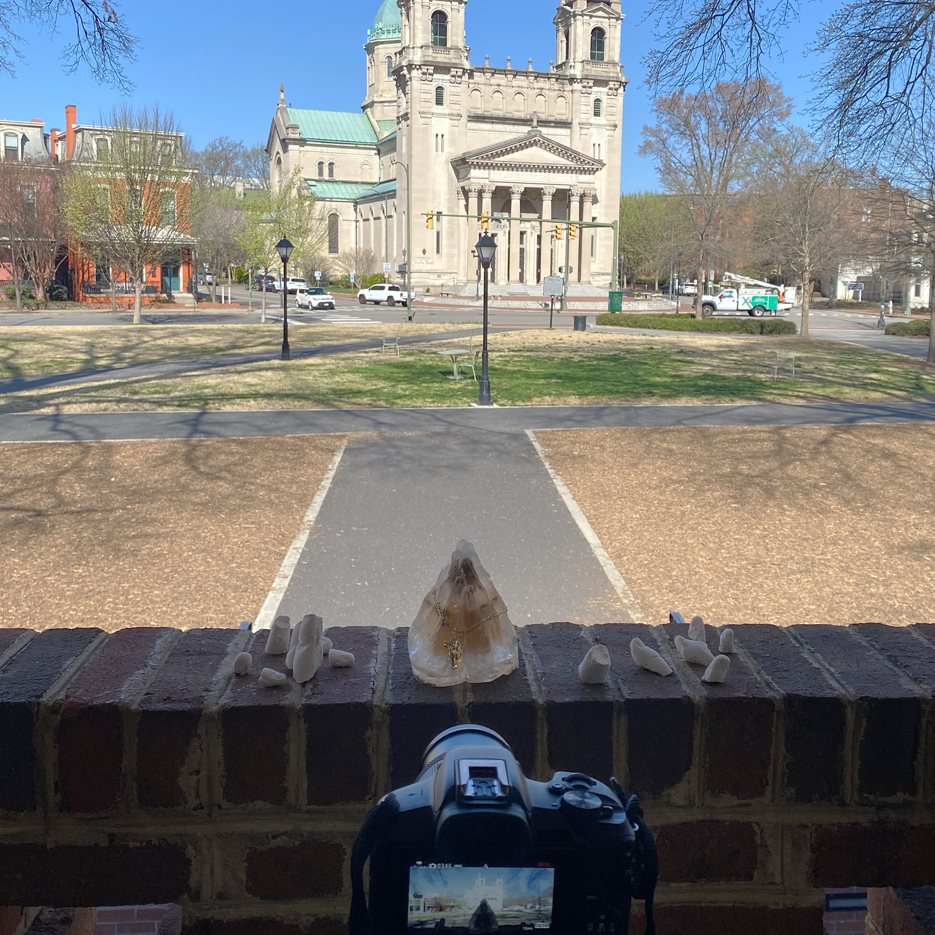 Image ID: A photograph of an ice sculpture of upright clasped hands on a ledge of brick overlooking a cathedral. On the ice sculpture is a gold necklace with a cross. Beside it are sculptures of plaster fingers. There is a camera focused on the ledge.